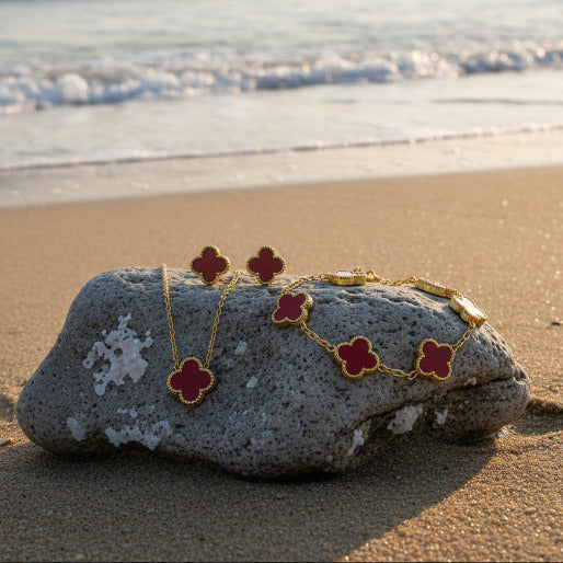 Gold jewelry set with red clover-shaped pendants including necklace, bracelet, and earrings on a beach background.