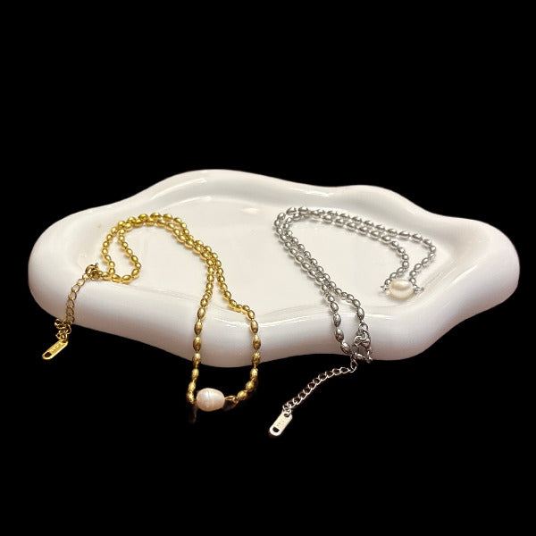 Two necklaces, one gold and one silver,  with beaded chains and a unique pearl as pendant, showed on a white dish with a black background. We can see the extender that gives versatility and flexibility.