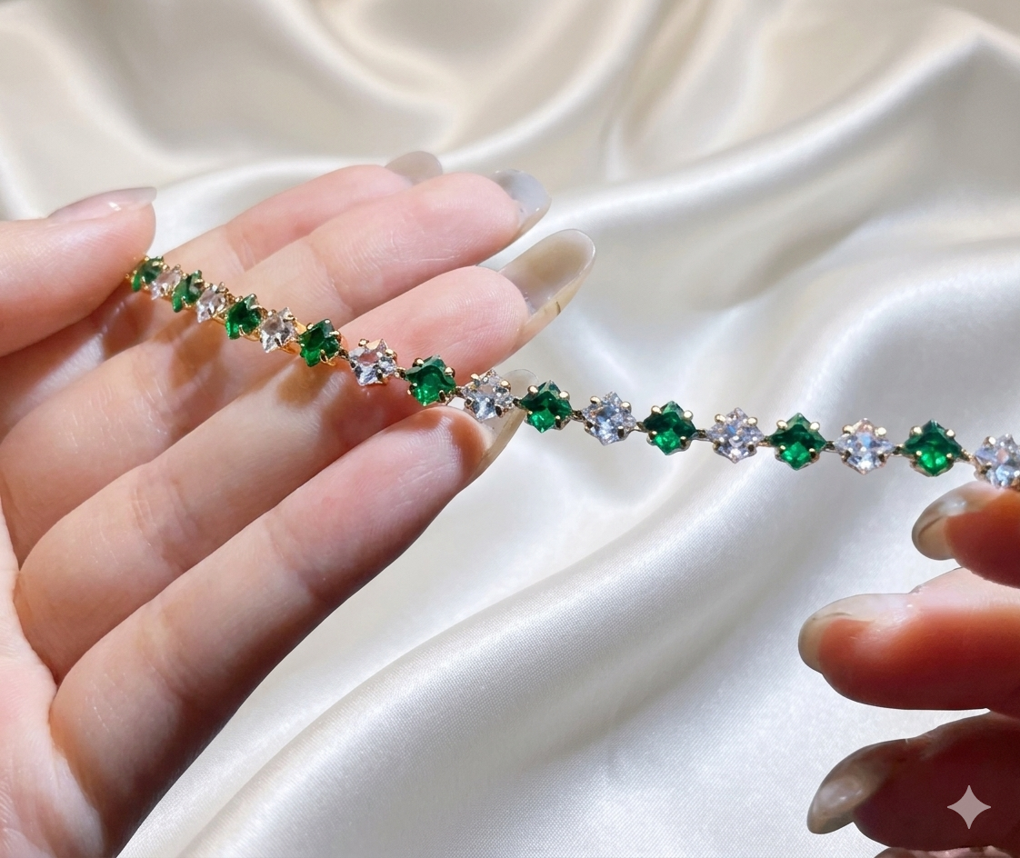 Tennis bracelet with green and clear gemstones held between two fingers on a white fabric background
