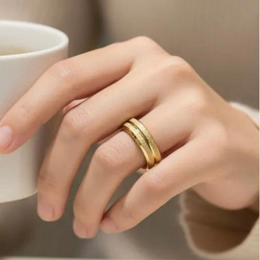 Hand wearing a gold ring holding a white mug. The most striking visual feature of this ring is its exquisite textured finish, Unlike a traditional polished band that reflects light directly, this granular surface diffused light, creating a soft, shimmering effect.