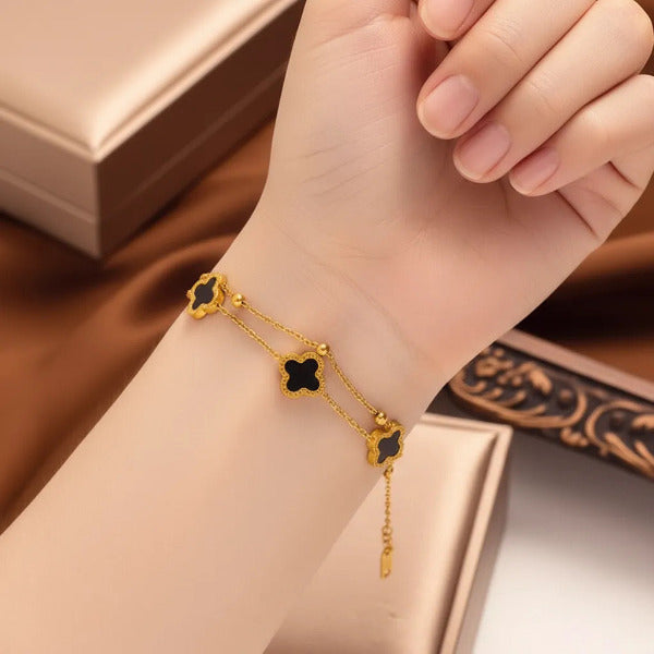 This is a delicate, gold tone double chain bracelet adorned with three distinct black clover-shaped charms. Each clover set is set with a thin gold border, adding a touch of elegance. Small spherical beads are interspersed along the chains.