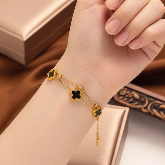 This is a delicate, gold tone double chain bracelet adorned with three distinct black clover-shaped charms. Each clover set is set with a thin gold border, adding a touch of elegance. Small spherical beads are interspersed along the chains.