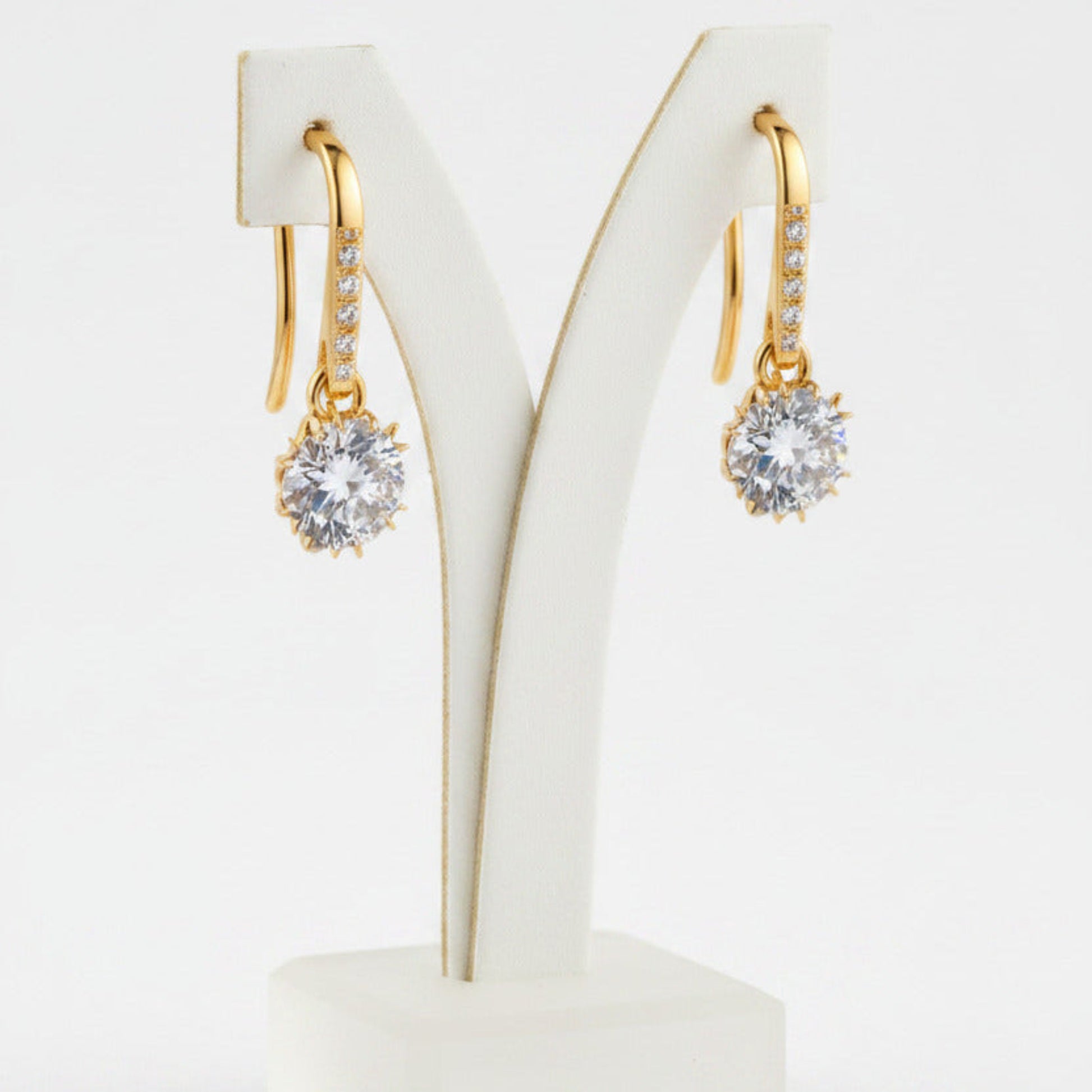 Gold earrings with clear moissanite gemstones on a white stand against a white background