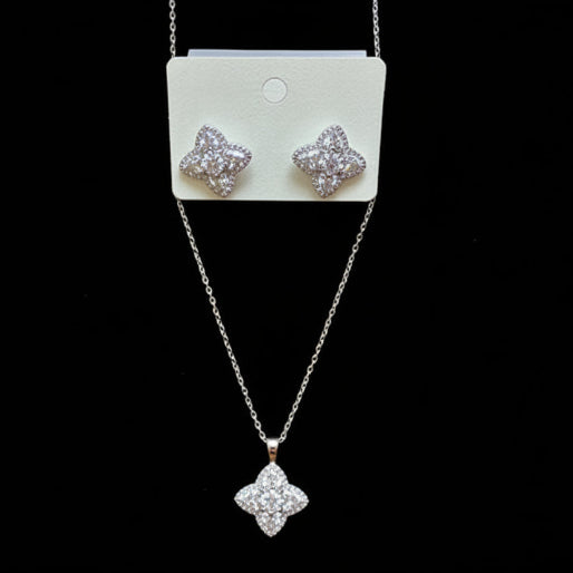 Designed with a four petal,  clover-like  motif, each petal has a subtle texture, and the entire shape is elegantly outlined with small, sparkling stones. A larger, clear, brilliant stoneis centrally placed in both the pendant and the earrings, adding a focal point to the design. Gold plated finished in gold tone or silver tone (like this one).