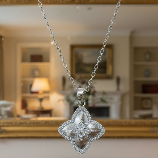 Detail of the necklace showing the symmetrical flower-shaped pendant adorned with stones. It is suspended from a delicate chain, secured with a spring ring clasp, and is designed to sit elegant;y around the neck as a statement or everyday accessory.