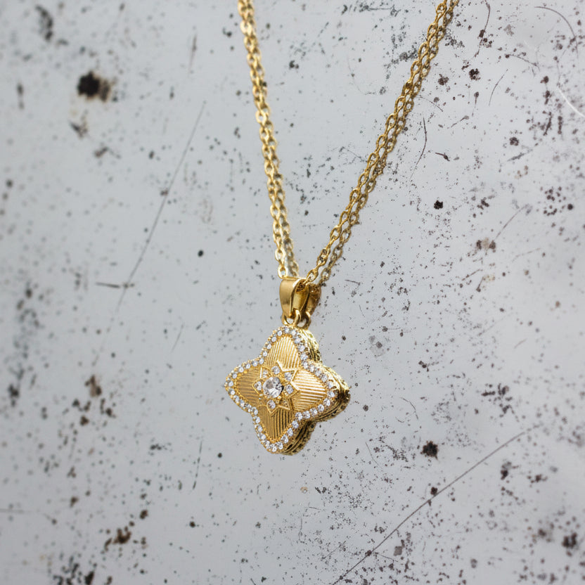 Detail of the necklace showing the symmetrical flower-shaped pendant adorned with stones. It is suspended from a delicate chain, secured with a spring ring clasp, and is designed to sit elegant;y around the neck as a statement or everyday accessory.