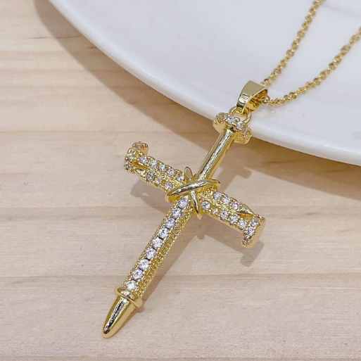 The pendant's design is formed by three stylized nails, two positioned horizontally to create the cross bar, and a third one forming the vertical beam. These 'nails' are intricately embellished with numerous small, sparkling clear stones, set in a pave fashion along their entire visible surface.