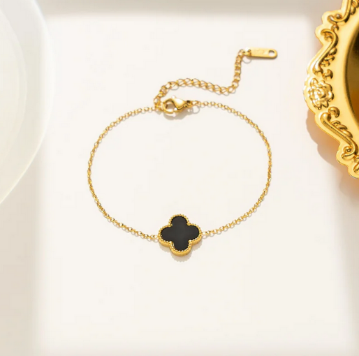 Gold bracelet with a black clover-shaped charm. Designed to be worn on the wrist, its classic aesthetic and adjustable length make it a versatile accessory, often symbolizing good luck and offering a touch of sophistication to any ensemble.