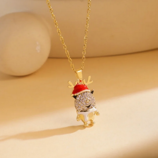 Christmas pendant necklace featuring a charming reindeer design, perfect for the holiday season. The reindeer is depicted wearing a red Santa hat with white trim and its body is entirely covered in small, sparkling clear stones set in a pave style. It hangs from a dainty gold-tone chain.